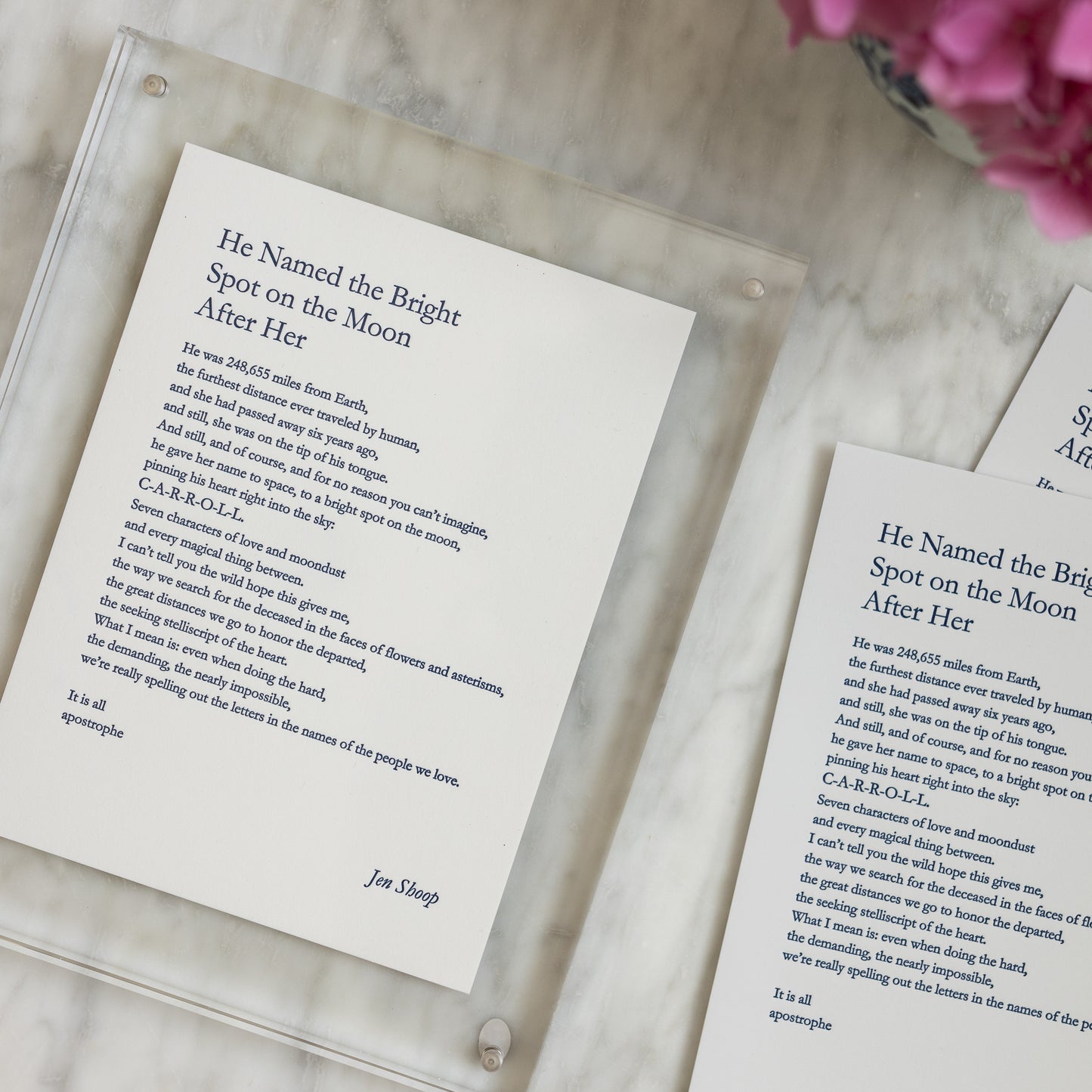 Styled flat lay of multiple “He Named the Bright Spot on the Moon After Her” letterpress prints with an acrylic frame and pink flowers.