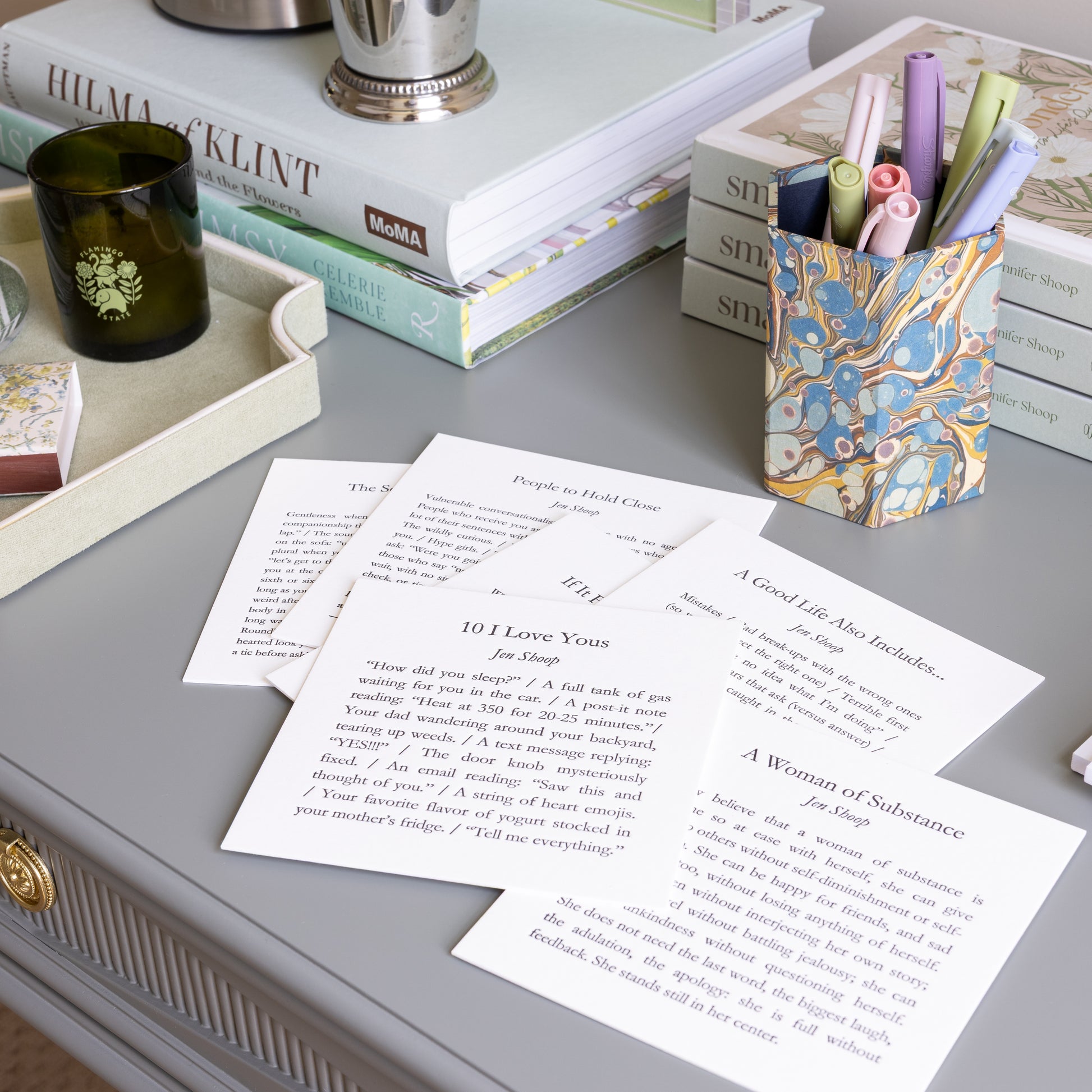 Six 5-by-5 poetry cards by Jen Shoop arranged across a styled writing desk with books, stationery and flowers.