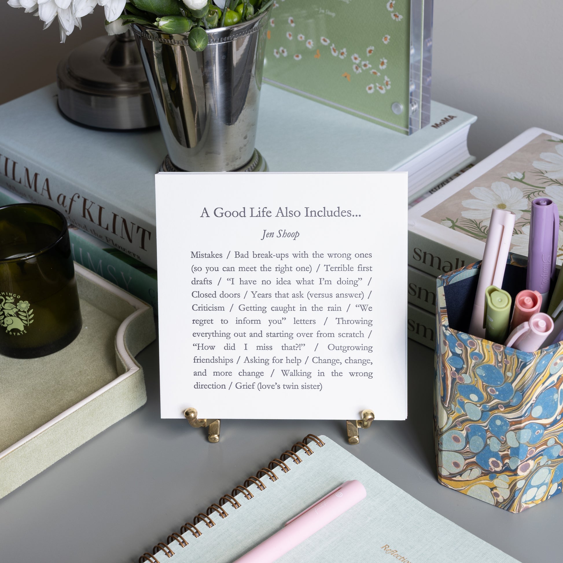 A Good Life Also Includes 5-by-5 poetry card by Jen Shoop displayed on a brass stand in a styled writing desk scene.