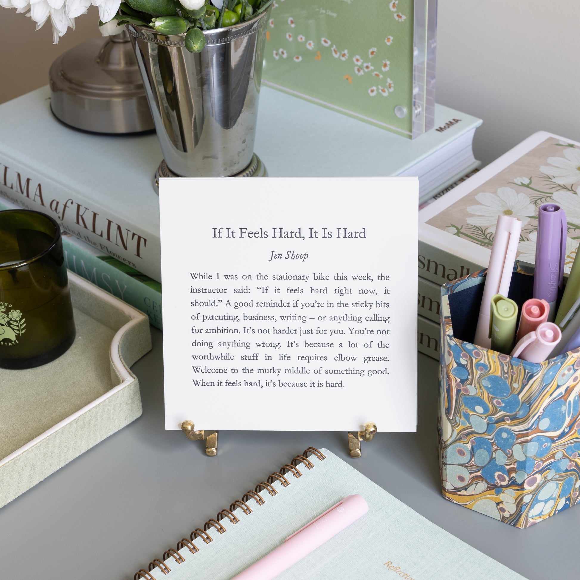 If It Feels Hard, It Is Hard 5-by-5 poetry card by Jen Shoop displayed on a brass stand in a styled writing desk scene.